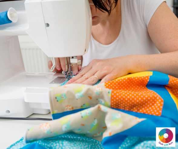 A woman quilting colorful, patterned fabric on a white sewing machine, focusing intently on her work; a Quilt with Kim logo appears in the bottom right corner.