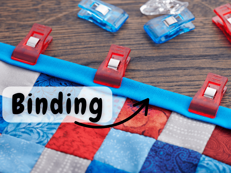 Close-up of a quilt edge with a bright blue fabric binding clipped in place using red and blue sewing clips. The quilt has red, blue, and grey patchwork squares. A label with the word 'Binding\' and an arrow points to the bound edge.