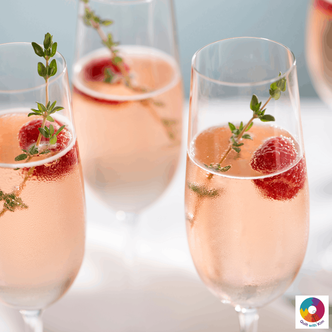 Four glasses of sparkling pink cocktail, each garnished with a sprig of thyme and a raspberry. The drinks are bubbly and served in champagne flutes.
