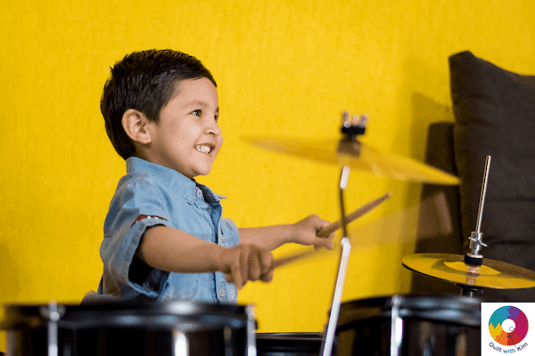 A young boy smiles while playing a drum set in front of a bright yellow wall. He holds drumsticks in both hands, appearing joyful and engaged in making music. A colorful logo is in the bottom right corner.