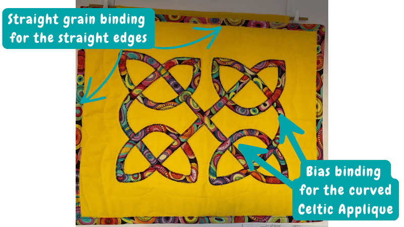 Photo of a yellow quilt with a red, purple, and teal Celtic knot appliqué design, annotated with arrows showing straight grain binding used on the straight outer edges and bias binding used on the curved appliqué shapes.