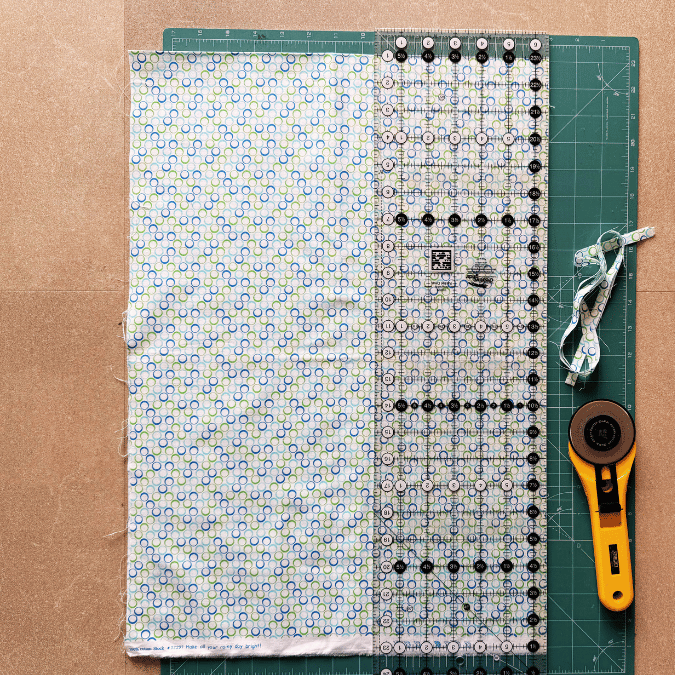 A patterned fabric, clear ruler, and rotary cutter are placed on a green cutting mat. Fabric strips are cut on the right side, with the ruler aligned along the fabric edge and trimmed pieces beside the rotary cutter.