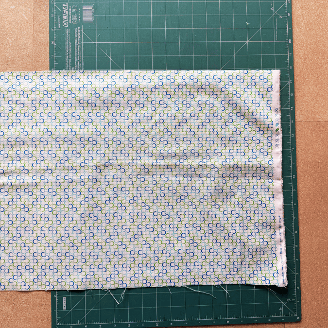 A piece of patterned fabric is folded and placed on top of a green cutting mat with measurement lines, on a brown tabletop.