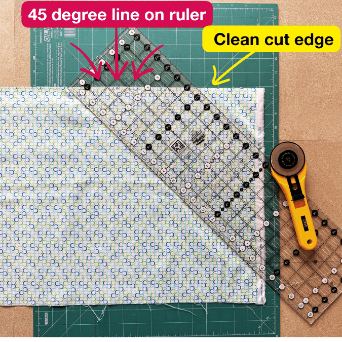 A quilting ruler is aligned at a 45-degree angle on patterned fabric on a cutting mat, with arrows showing the 45-degree line and a label pointing to the clean cut edge. A rotary cutter lies nearby.