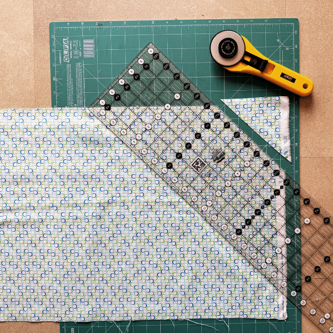 A rotary cutter, quilting ruler, and green cutting mat are shown with a piece of patterned fabric being measured and cut, with a small triangle already trimmed.