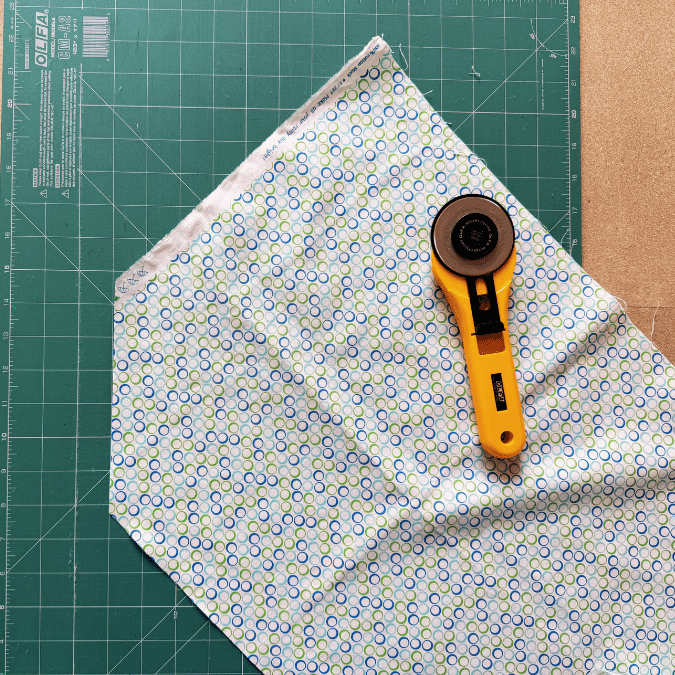 A yellow rotary cutter rests on patterned fabric with blue and green circles, placed diagonally on a green cutting mat with measurement lines. Part of a brown surface is visible in the corner.