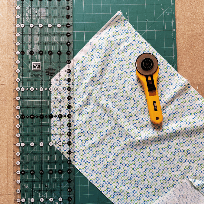 A rotary cutter, a clear quilting ruler, and printed fabric on a green cutting mat, all prepared for fabric cutting and sewing projects.