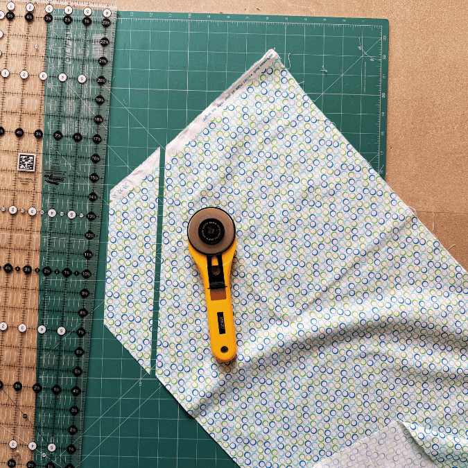 A rotary cutter, ruler, and patterned fabric are on a green cutting mat. The fabric is partially cut, showing precise measurement and alignment for sewing or quilting.