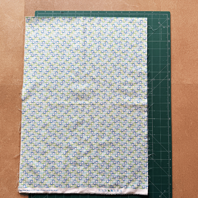A patterned fabric with small circular designs is laid on top of a green cutting mat with measurement markings, placed on a brown surface. The fabric mostly covers the mat, revealing its grid on one side.