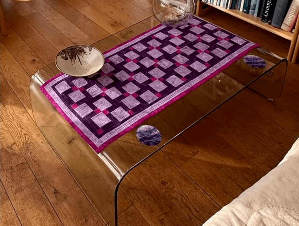 Purple and pink patchwork table runner made from geometric quilt blocks, displayed on a glass coffee table.