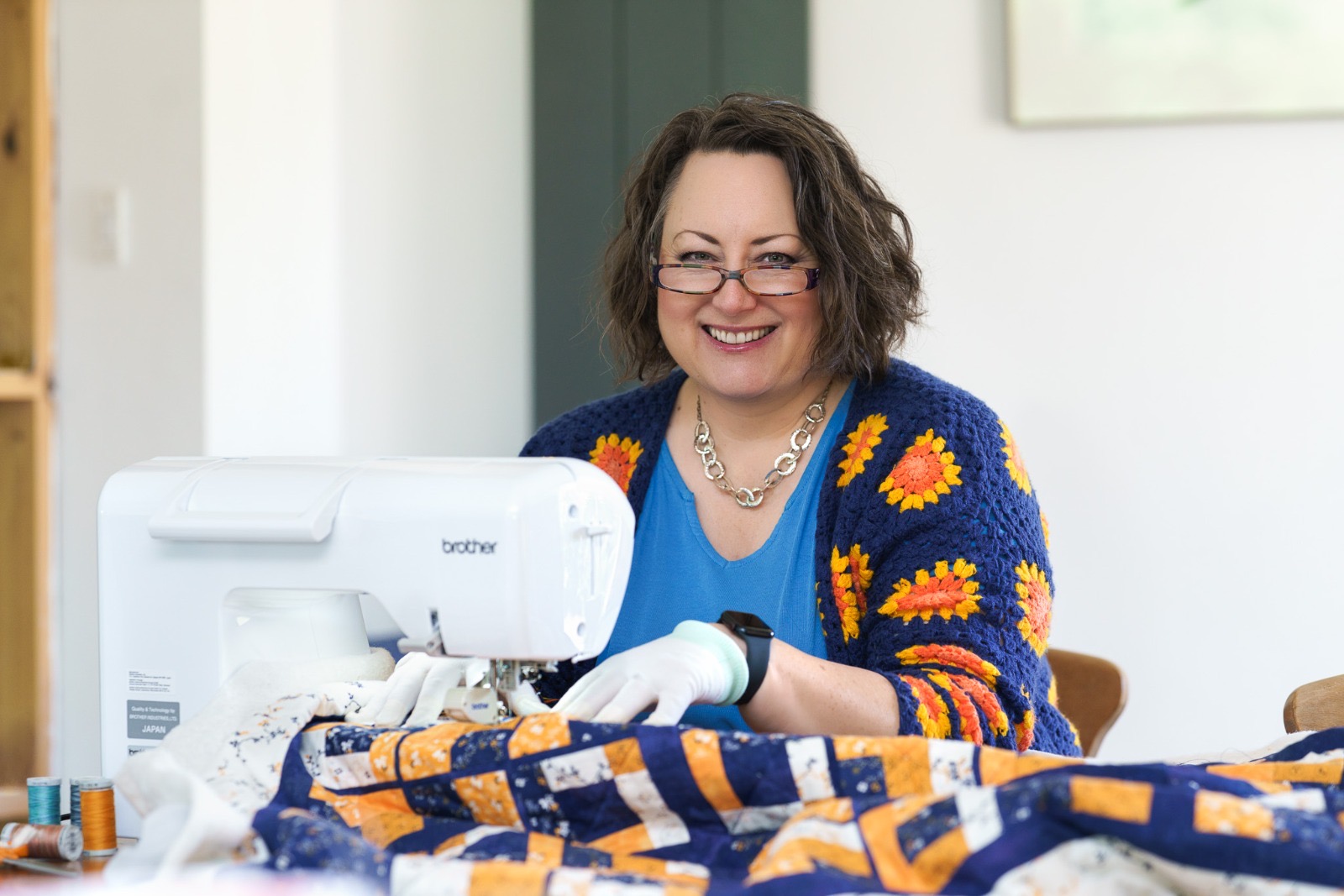 A smiling woman with short gray hair and glasses on her head is wearing a teal lace top and a smart watch, sitting in a colorful room decorated with quilts and crafts.